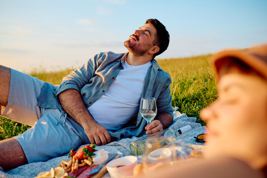 Man relaxing on a picnic blanket, soaking up the warm sun with eyes closed, holding a wine glass in a picturesque field