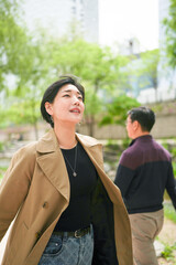 Naklejka premium Woman smiles softly, eyes upward, in urban park. Brown coat, black top, and jeans complete her casual look. Background shows a man walking away, blurred by depth