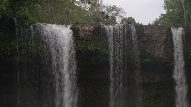 Static shot of Tad Champee waterfall with multiple curtains of water falling over a rocky ledge into a jungle pool in Laos.