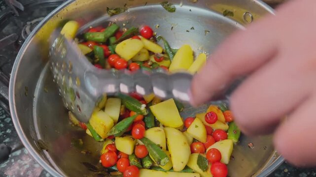 Close view of fresh okra potatoes and cherry tomatoes being stirred inside a steel cooking pan with visible spices and herbs during homemade vegetable preparation in a kitchen environment