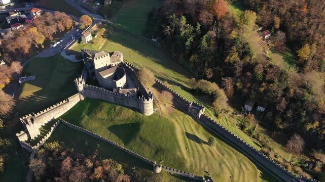 Aerial view of Castel Grande with its medieval stone walls and towers surrounded by autumn trees and green hills in Bellinzona, Ticino, Switzerland.