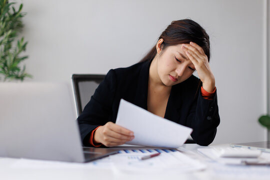 Asian businesswoman holding a document, suffering from a headache and work stress in an office, facing financial problems, debt, and burnout from demanding tasks and a tight deadline