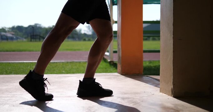 Cinematic low angle close-up of a male athlete's legs and feet doing a standing calf stretch on concrete at an outdoor sports stadium