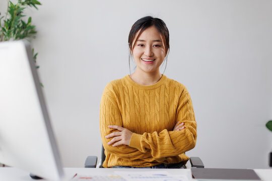 Asian professional woman sitting at her modern office desk, smiling confidently with arms crossed, projecting success, creativity and career ambition in a bright work environment