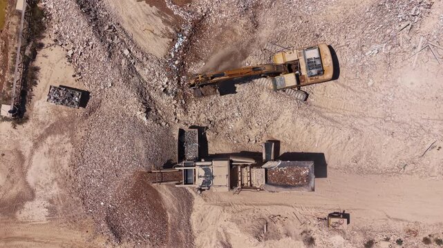 Overhead view of a yellow excavator processing construction rubble and debris at a demolition site, with a crusher machine sorting materials on sandy ground