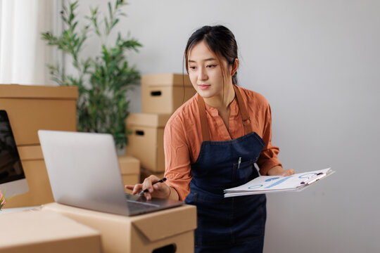 Asian female business owner managing online store operations, checking orders and inventory on laptop while preparing packages for e-commerce shipping