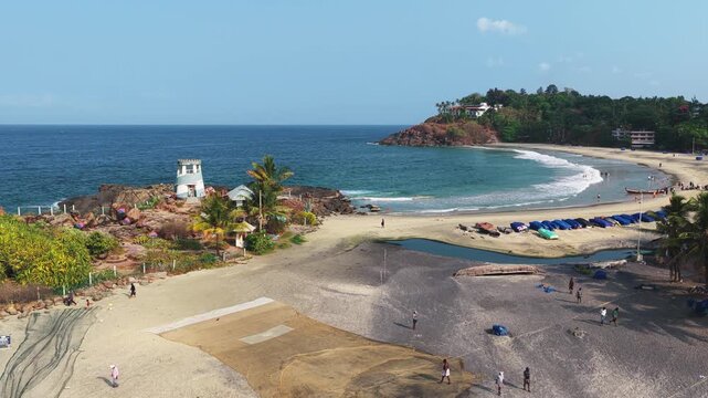 Aerial view of the sandy beach with fishing nets, a small white tower on the rocks, and people walking along the shore in Kovalam, Kerala, India.