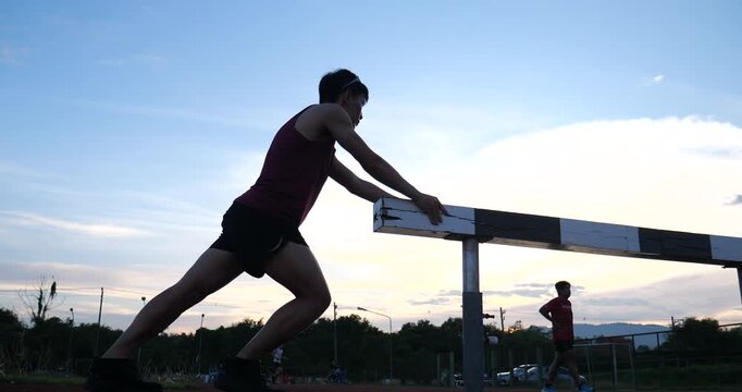 Cinematic low angle shot of a male runner doing a calf stretch against a steeplechase hurdle on track during twilight sunset, athletic conditioning concept