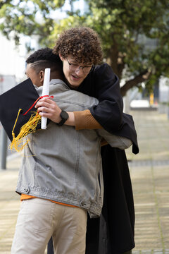 Diverse males hugging on path, graduate in gown holding diploma and cap, friend in denim jacket