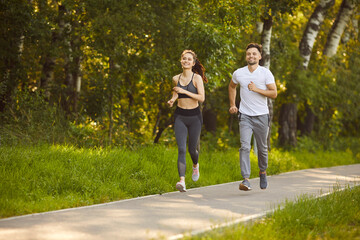 Obraz premium Happy athletic couple jogging together in park path surrounded by green trees. Smiling sporty young man and woman enjoying outdoor run on sunny day, staying fit and active. Healthy lifestyle concept.
