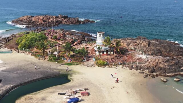 Aerial view of a white lighthouse on a rocky outcrop surrounded by palm trees and people walking on the sandy beach in Kovalam, Kerala, India.