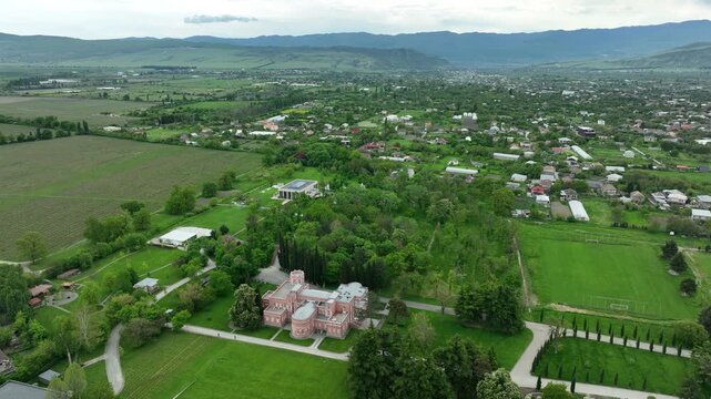 Aerial view of Chateau Mukhrani royal palace and estate in Georgia, magnificent pink castle architecture with a tower surrounded by a lush green park, gardens and pathways on a spring day.