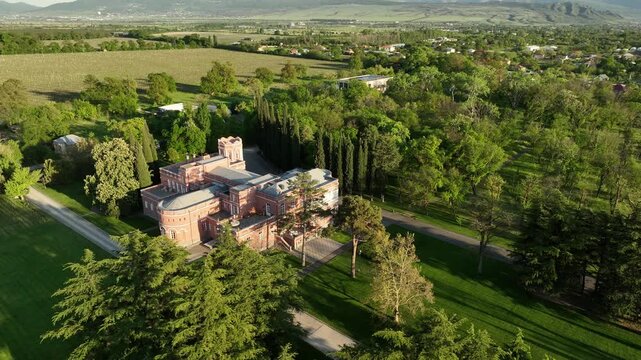 Aerial view of Chateau Mukhrani royal palace and estate in Georgia, magnificent pink castle architecture with a tower surrounded by a lush green park, gardens and pathways on a spring day.