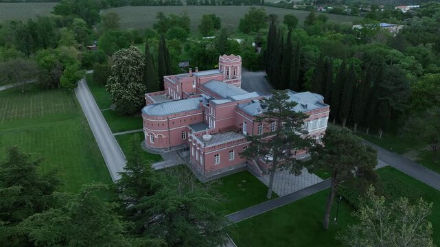 Aerial view of Chateau Mukhrani royal palace and estate in Georgia, magnificent pink castle architecture with a tower surrounded by a lush green park, gardens and pathways on a spring day.
