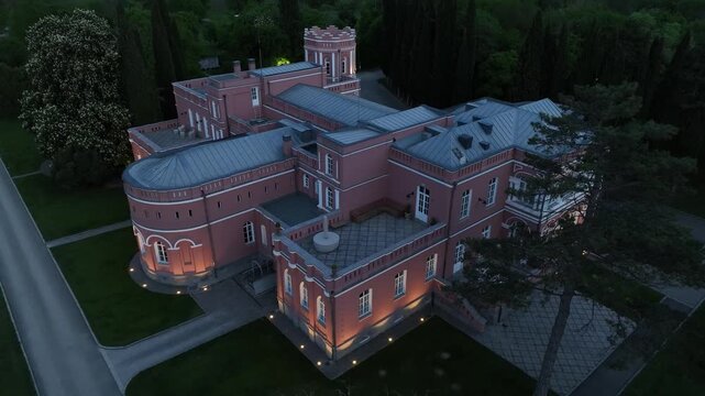 Aerial view of Chateau Mukhrani royal palace and estate in Georgia, magnificent pink castle architecture with a tower surrounded by a lush green park, gardens and pathways on a spring day.