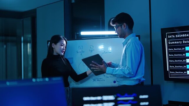 Professional software engineers, a man and woman, collaborate on a coding flow whiteboard while analyzing big data dashboards on screens in a dark, high-tech IT development office late at night.