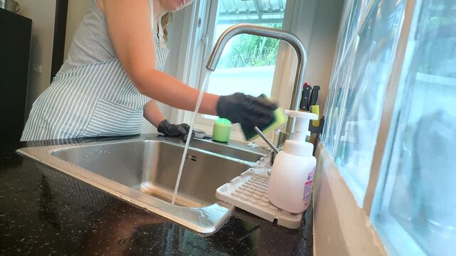 Woman cleaning kitchen sink with sponge under running water while wearing black rubber gloves. Real life video footage of home cleaning, washing sink, kitchen hygiene and everyday household chores.