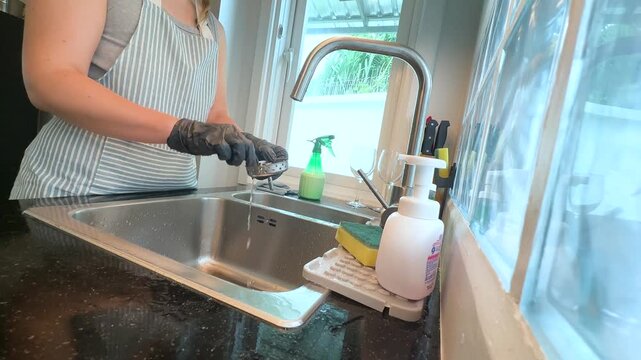 Woman cleaning sink drain strainer by hand while wearing black rubber gloves in kitchen. Real life video footage of home cleaning, sink maintenance, kitchen hygiene and everyday household chores.