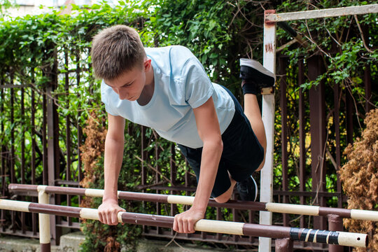 Teenage boy practicing challenging calisthenics skill on outdoor parallel bars, focused adolescent athlete maintaining static position and developing strength, coordination and fitness through bodywei