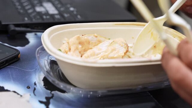 a person eating a chicken rice bowl with sauce at a work desk. A laptop and smartphone are visible in the background, depicting a busy office lunch break or remote work lifestyle.
