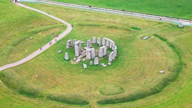 Close aerial drone footage of Stonehenge in Wiltshire, England, showing the prehistoric standing stones, ancient stone circle, surrounding earthworks, visitor paths, and green grassland.