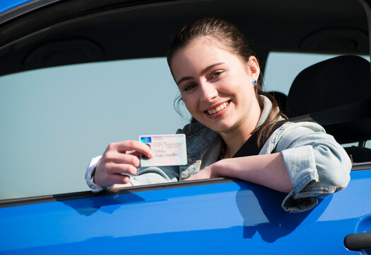Smiling teenage girl sitting in car showing driving license, partial view