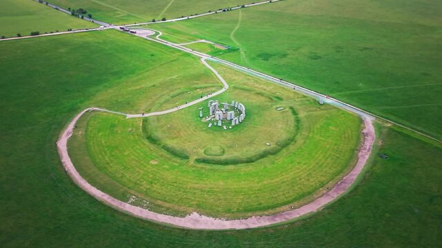 Aerial drone footage of Stonehenge in Wiltshire, England, showing the ancient stone circle surrounded by green grassland, pathways, visitor access routes, and the wider rural landscape.