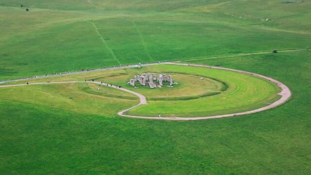 Wide aerial drone footage of Stonehenge in Wiltshire, England, showing the ancient prehistoric stone circle surrounded by circular earthworks, visitor paths, tourists, and open green grassland across