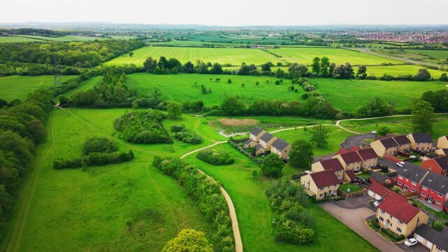 Aerial drone footage showing suburban houses on the edge of green countryside, with farmland, hedgerows, walking paths, trees, ponds, roads, and open fields visible beyond the residential area.