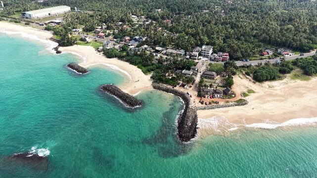 Aerial view of the tropical coastline featuring sandy beaches, rock breakwaters, and lush palm trees along the shore in Peraliya, Southern Province, Sri Lanka.