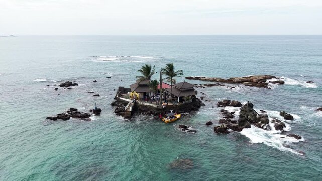 Aerial view of Seenigama Muhudu Dewalaya temple on a rocky island with palm trees and a small boat in the turquoise ocean Peraliya, Sri Lanka.