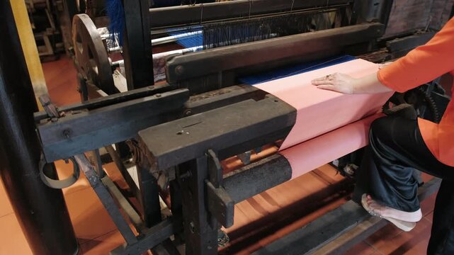 Woman working at traditional wooden loom to weave pink silk fabric in textile factory, Vietnam