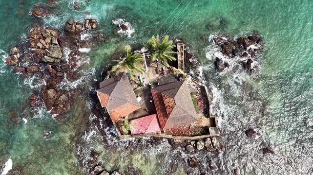 Aerial view of Seenigama Muhudu Dewalaya temple on a rocky island with palm trees and turquoise waves crashing against the shore, Peraliya, Sri Lanka.