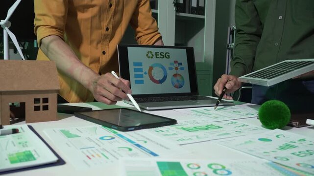 Team reviewing esg reports and green business charts on a desk with laptop, tablet and model house in an office, showing sustainability planning and eco strategy.