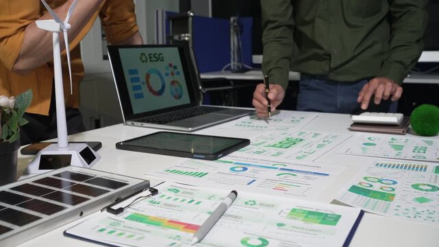 Team reviewing es g charts and sustainability reports at an office desk with laptop, tablet, solar panel and wind turbine model, showing green business planning and renewable energy goals.