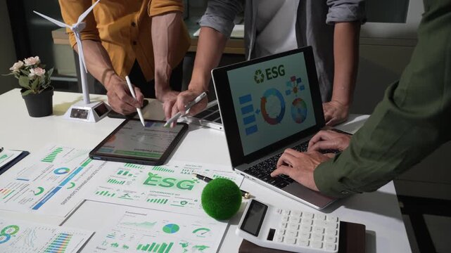 Team reviewing esg charts and sustainability data on laptops and papers during an office meeting, with a wind turbine model and calculator on the desk