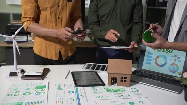 Team reviewing sustainability reports at an office desk with charts, laptop, tablet, model house and small wind turbine, showing green business planning and environmental goals.