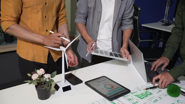 Team meeting around a desk with wind turbine model, tablet and charts, discussing renewable energy, sustainability and es g planning in a modern office