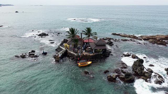 Aerial view of Seenigama Muhudu Dewalaya temple on a rocky island with palm trees and a yellow boat in the ocean Peraliya, Sri Lanka.
