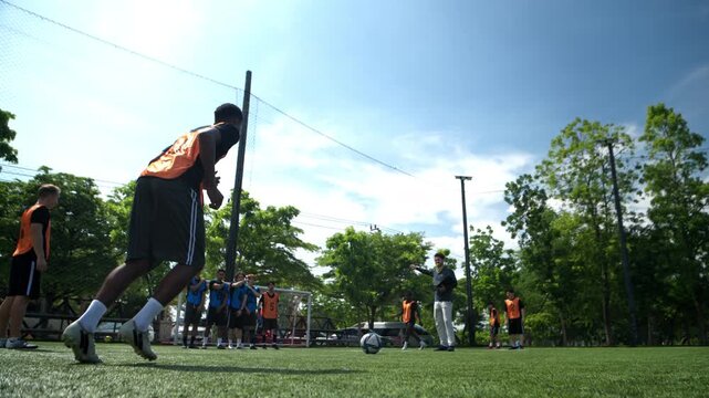 A young football players are practicing free kicks while the opposing team forms a defensive wall, with a coach giving them instructions on an outdoor football field