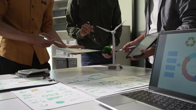 Business team reviewing charts around a model wind turbine and recycling report in an office, showing planning, teamwork and clean energy strategy.
