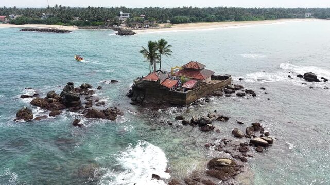Aerial view of Seenigama Muhudu Dewalaya temple on a small rocky island surrounded by turquoise ocean waves and palm trees near the coast Peraliya, Sri Lanka.