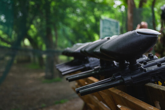 Row of paintball marker guns arranged on wooden rack at outdoor field. Protective equipment prepared for recreational team competition and active leisure activities.