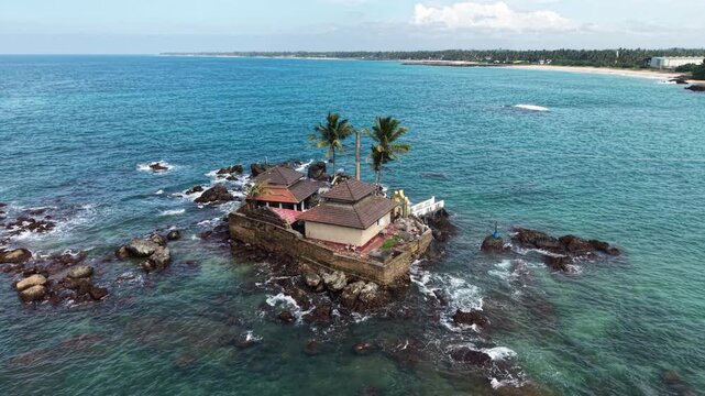 Aerial view of Seenigama Muhudu Dewalaya temple on a rocky island surrounded by turquoise ocean waves and palm trees in Peraliya, Sri Lanka.