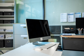 Naklejka premium Modern office desk with desktop computer, keyboard and mouse in a clean workspace. Glass wall, shelves and filing cabinet create a professional business setting.