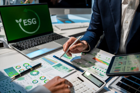 Business team reviews esg reports and green finance charts at office desk with laptop, tablet and calculator, showing sustainability planning, audit work and corporate reporting.