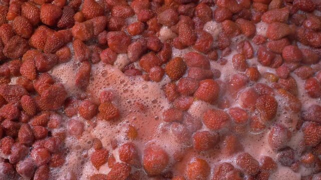 Strawberries are boiling in a big vessel, steam rising as they bubble. The thick mixture is continuously stirred with a spoon to keep it from sticking and to mix the ingredients