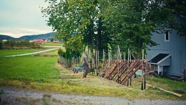 A Person is Building a Wooden Fence Along the Side of a Property, Surrounded by Green Fields and Trees - Timelapse
