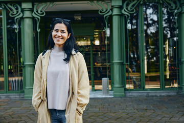 Obraz premium Woman in beige coat and gray sweater stands outdoors in front of a green storefront, smiling in an urban daylight scene, blending casual fashion with a relaxed city mood and bright daytime ambiance.