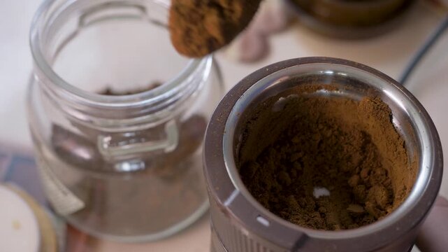 Hand fills container with fresh coffee powder from grinder. Glass jar holds aroma meant to energize mornings and improve focus. Roasted coffee beans have been ground down to be brewed for coffee